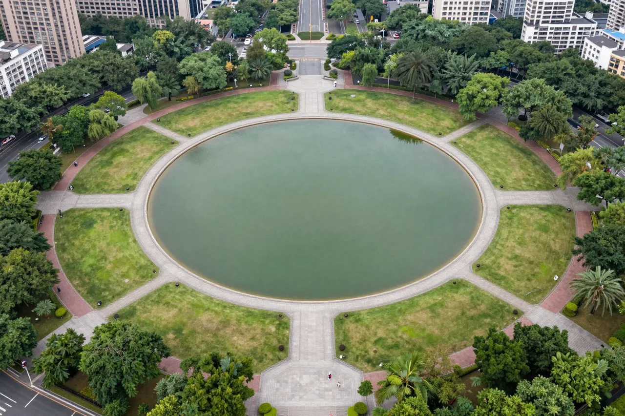 Overhead aerial view of a formal urban park centered on a round pond with radiating paved paths and surrounding city
