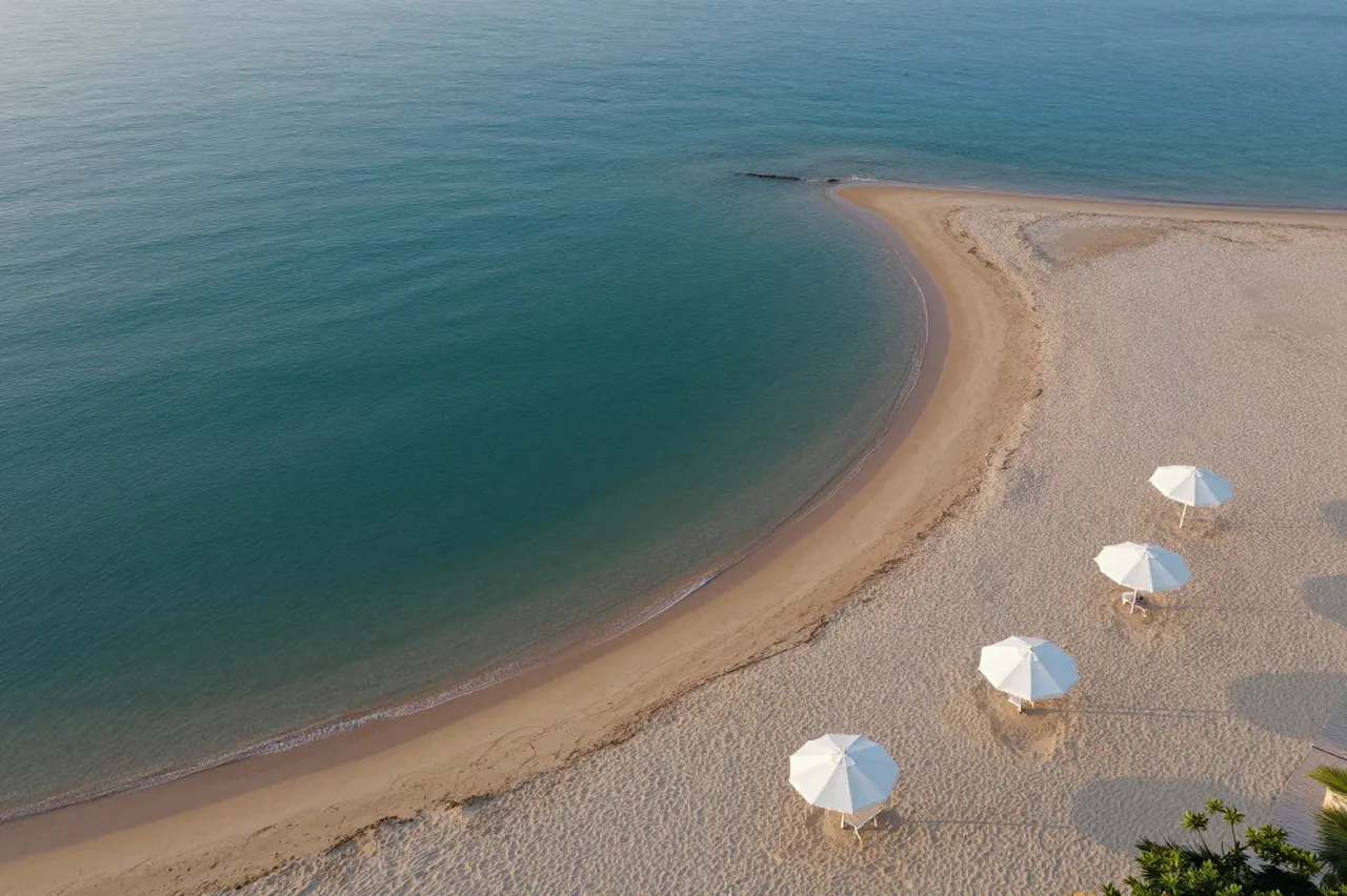 Aerial perspective of a calm crescent beach lined with white umbrellas next to a turquoise bay at early morning.