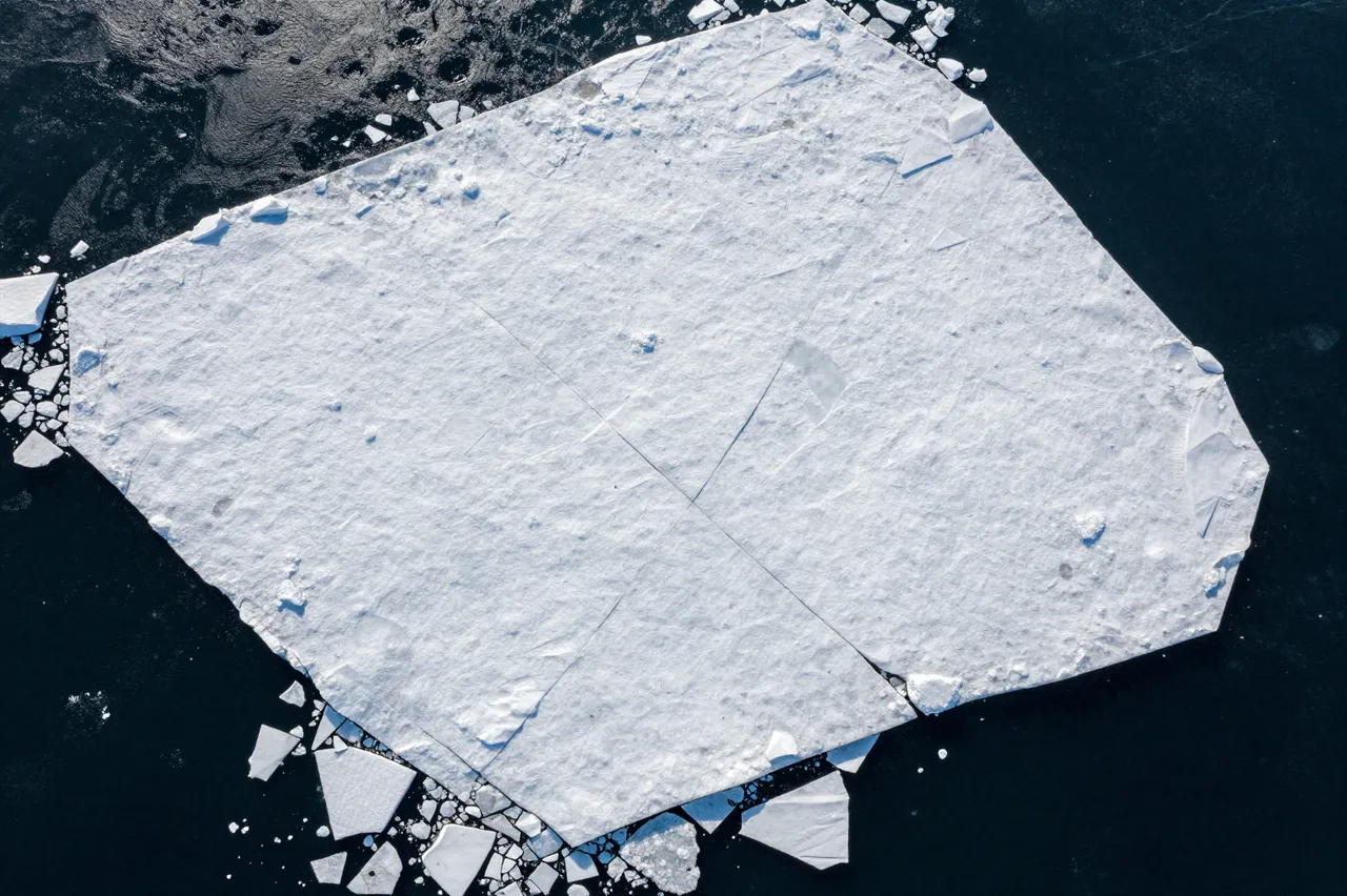 Top-down aerial view of a large flat drift ice plate floating on dark open ocean water with surrounding smaller ice fragments