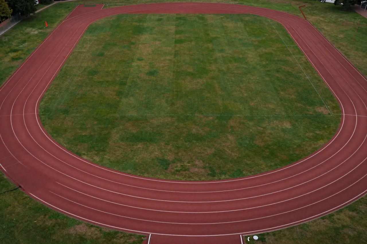 Top-down aerial view of an empty red running track surrounding a grass sports field at a recreation ground.