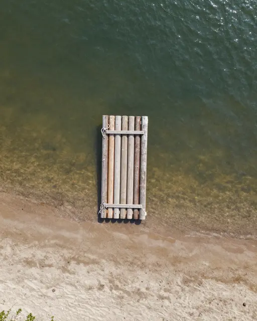 A top-down aerial shot of a wooden log raft floating near a sandy lakeshore