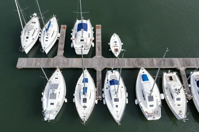 Top-down aerial view of white sailboats moored to wooden piers on green marina water.