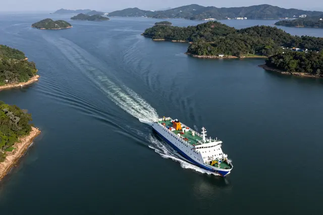 A high angle view of a passenger ferry moving through dark blue water between forested green islands, leaving a curved wake