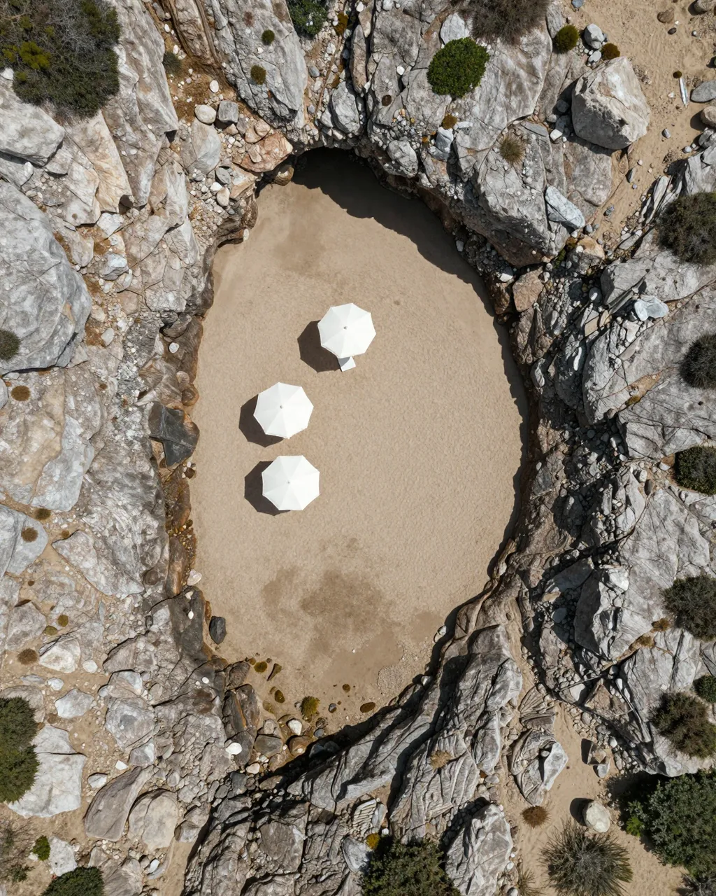 Top-down aerial shot of a tiny sandy rocky cove with three white beach umbrellas under bright sun.