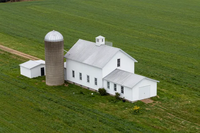 Aerial shot of a white farm building, concrete silo and outbuildings surrounded by green crop fields.