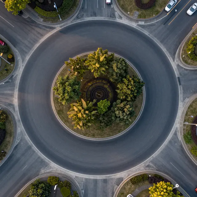Top-down aerial view of a paved roundabout with a tree-filled center at dawn, with a few parked cars on approach roads.
