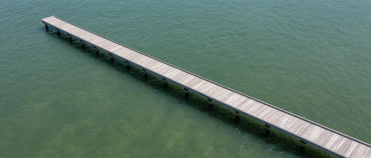 High angle aerial view of a long wooden pier stretching across calm green clear water