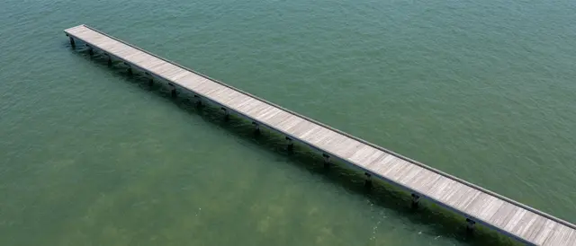 High angle aerial view of a long wooden pier stretching across calm green clear water