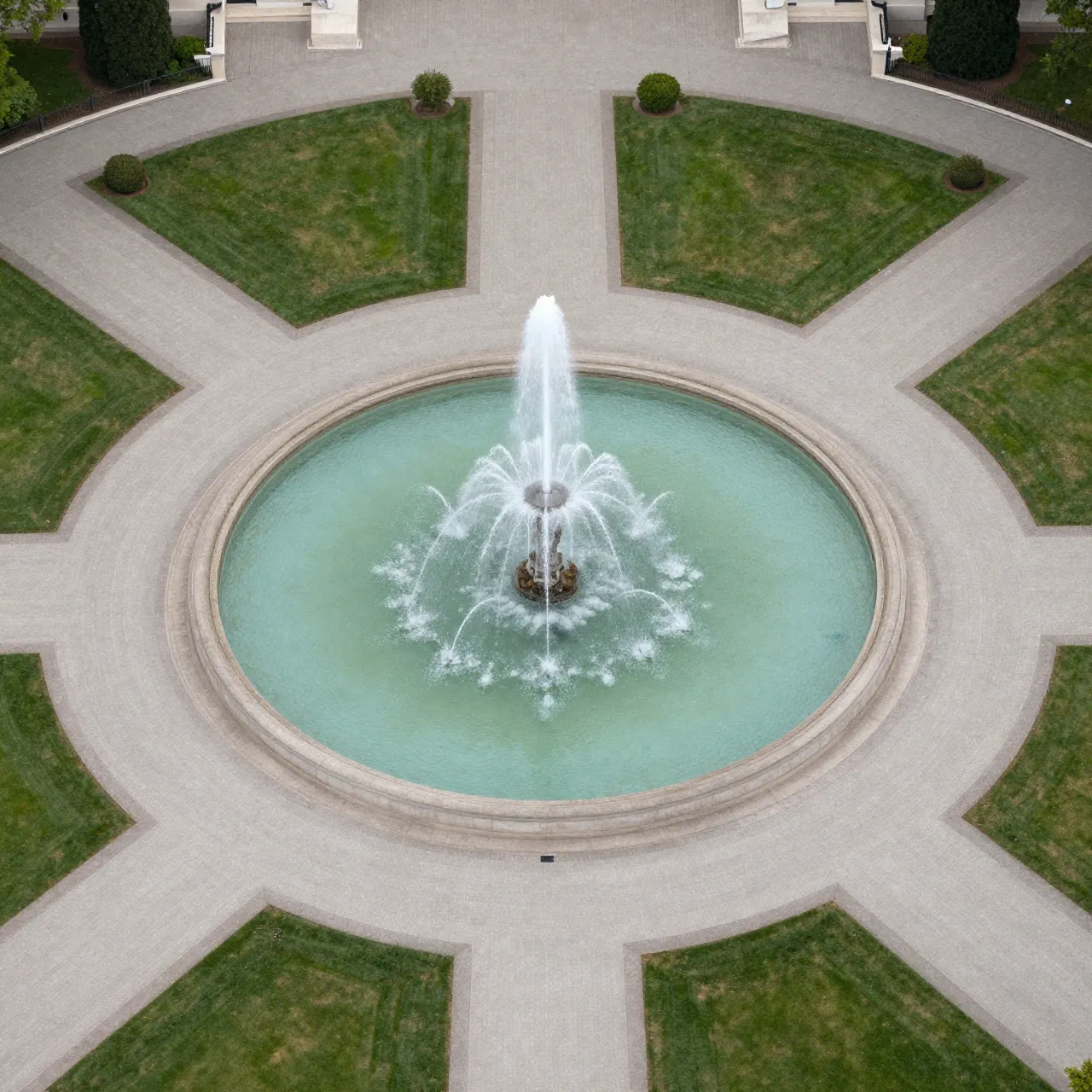 High-angle aerial view of a circular central fountain with radiating paved paths and symmetric lawns.