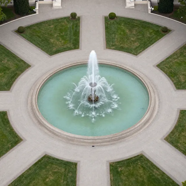 High-angle aerial view of a circular central fountain with radiating paved paths and symmetric lawns.