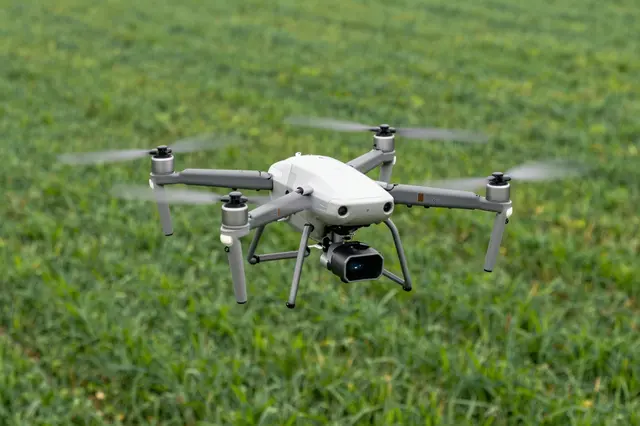A quadcopter drone hovers mid-air above a lush green farm field with spinning blurred propellers