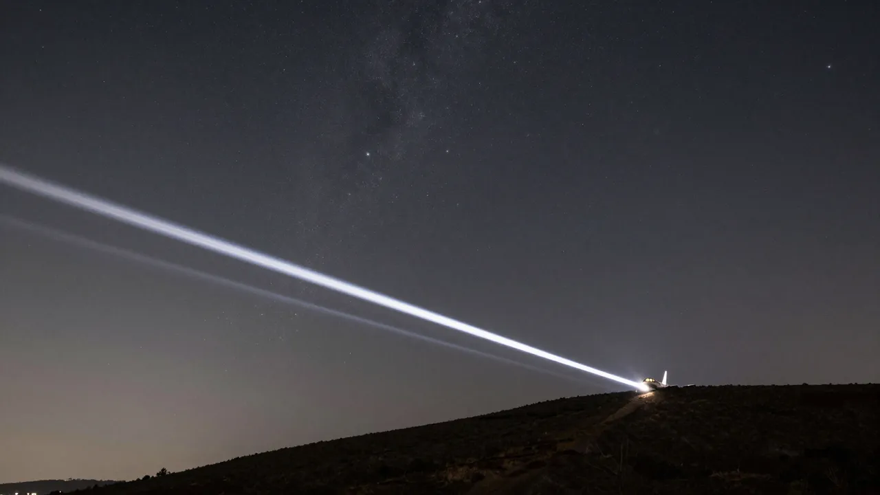 A long bright aircraft light streak crosses a starry night sky above a dark sloped hillside