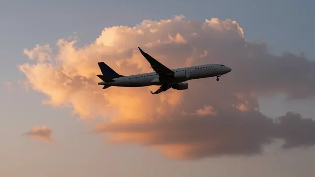 A passenger airliner flies with extended landing gear in front of a large orange-pink lit cumulus cloud at sunset