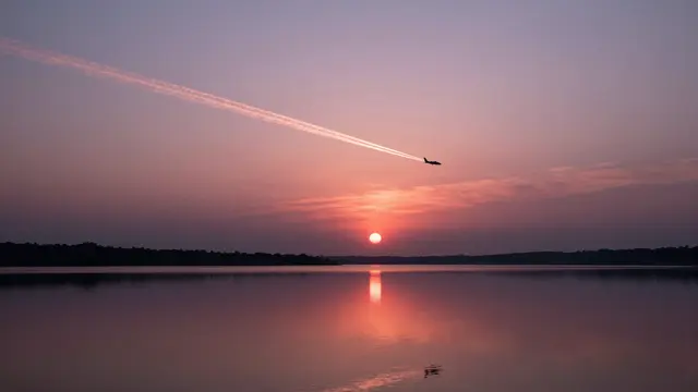 A tiny airliner with a glowing contrail flies over a still pink and purple sunset lake