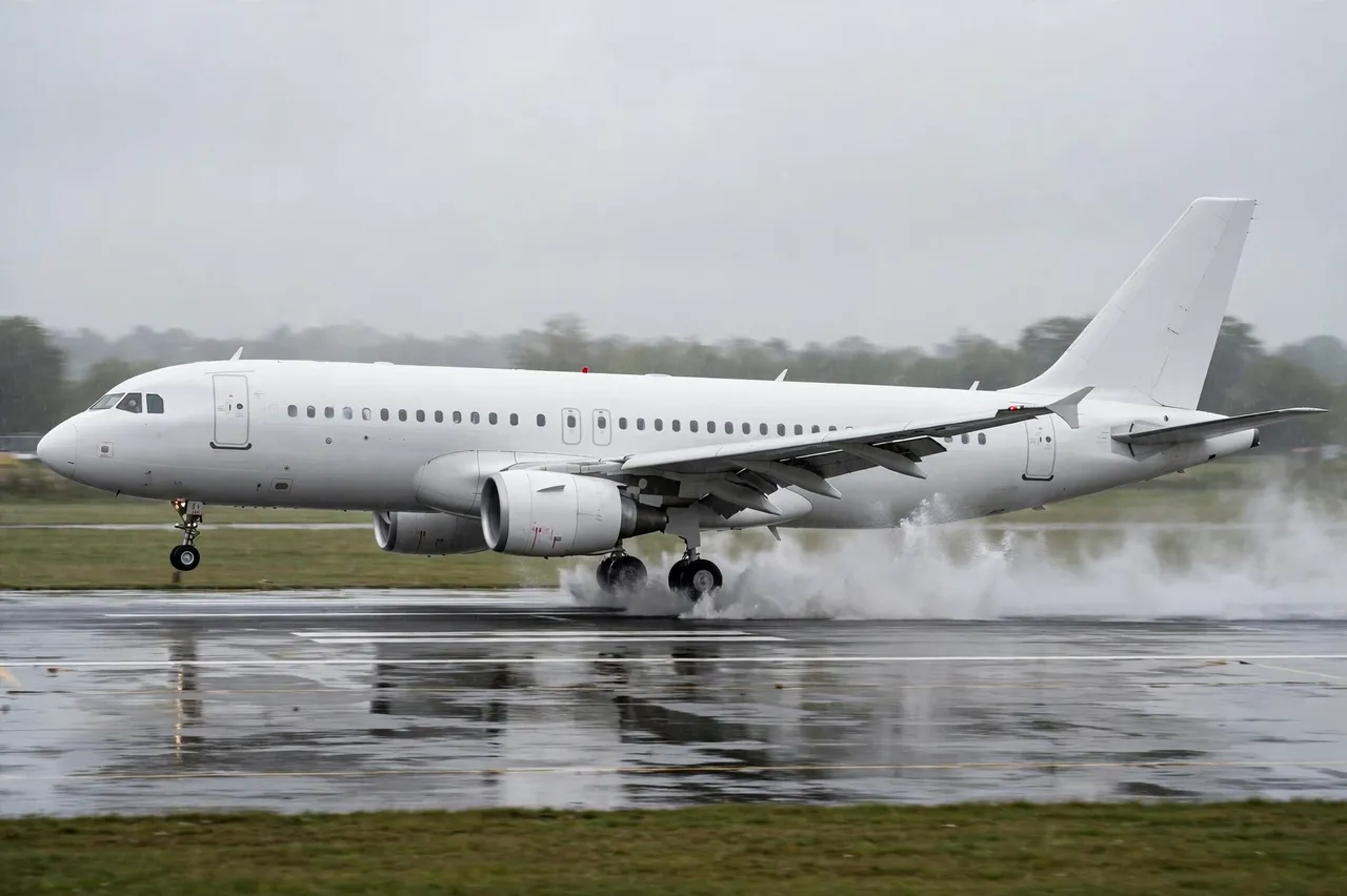 Plain white modern airliner touches down on a wet airport runway, kicking up water spray from its landing gear in side view.