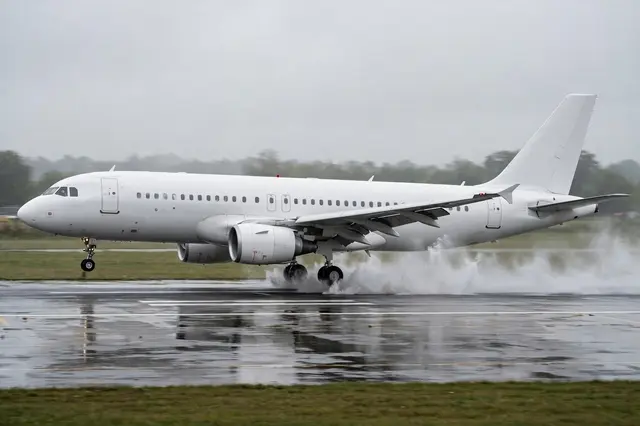 Plain white modern airliner touches down on a wet airport runway, kicking up water spray from its landing gear in side view.