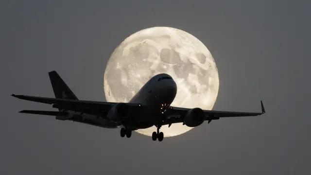 Silhouetted commercial jet with landing gear extended flies directly in front of a large bright full moon