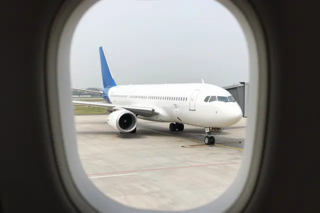 Unbranded white airplane with blue tail sits on airport tarmac seen through an aircraft window