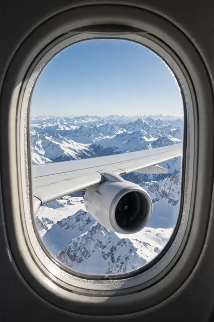 View through an oval airplane cabin window showing the plane wing, engine, and snow covered mountains under a clear blue sky