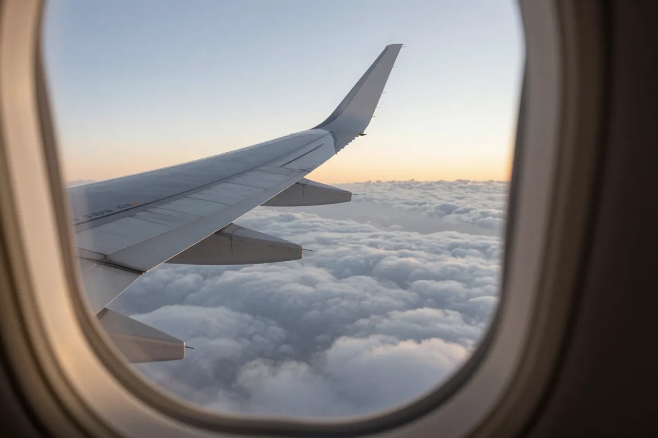 View from an airplane cabin window of the wing floating above fluffy clouds during soft sunset