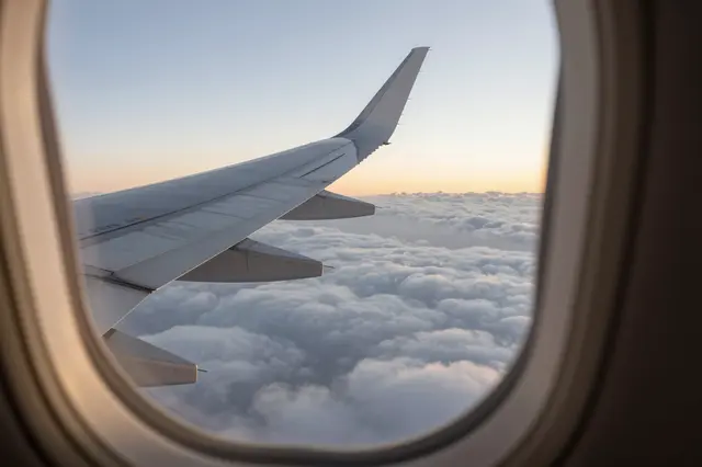 View from an airplane cabin window of the wing floating above fluffy clouds during soft sunset