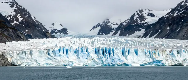 A jagged blue and white tidewater glacier meets calm ocean water beneath snow-capped Alaska mountains under an overcast sky
