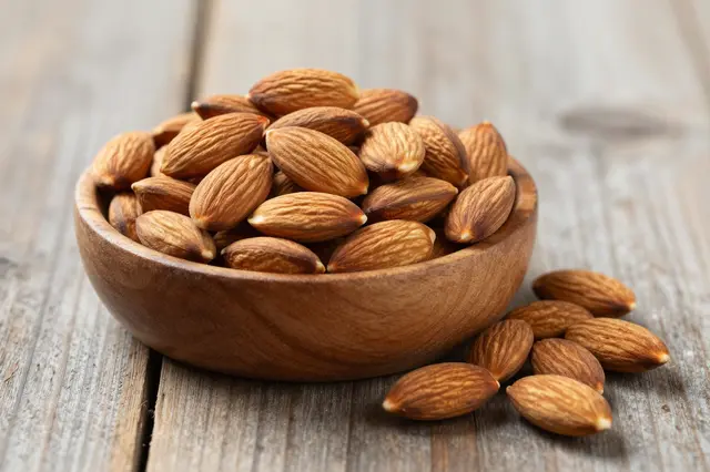 A wooden bowl filled with whole almonds sits on a weathered wooden table with several almonds spilled beside it.