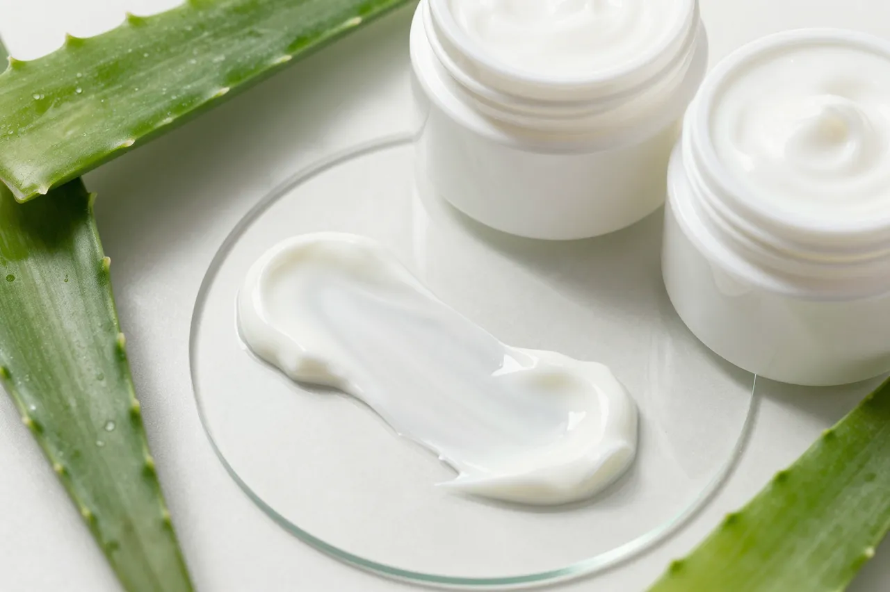 Creamy skincare swatch sits on a glass disc beside open white jars and fresh aloe vera leaves.