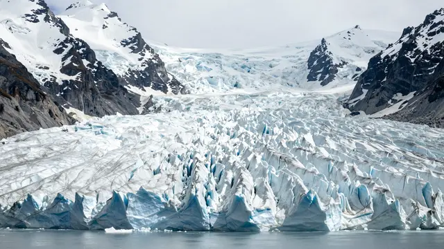 A jagged blue and white glacier flows between rocky snow-capped mountains down to calm glacial water