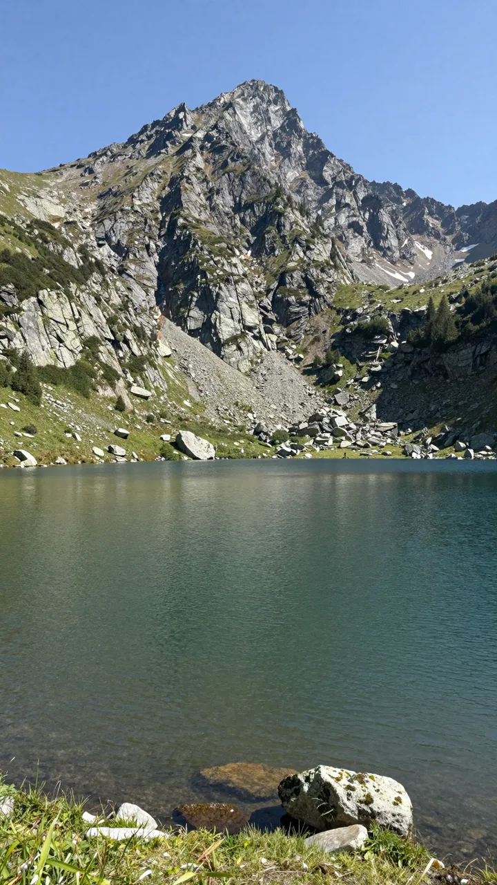 Clear turquoise mountain lake sits below a jagged rocky peak under a bright summer blue sky