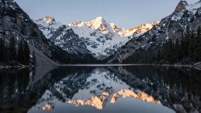 Snowy jagged mountain peaks are perfectly mirrored in a still calm lake at early dawn