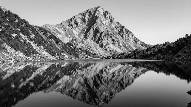 A black and white view of a calm glassy alpine lake reflecting a sharp steep mountain peak