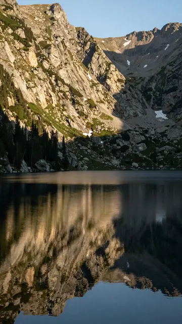 Calm mountain lake reflects sunlit rocky alpine peaks and forest under a clear pale sky at sunrise