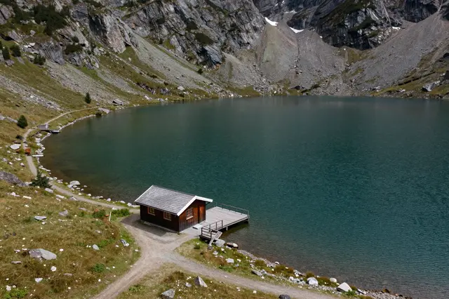 Aerial view of a wooden cabin with dock on the shore of a turquoise alpine lake surrounded by rocky mountains