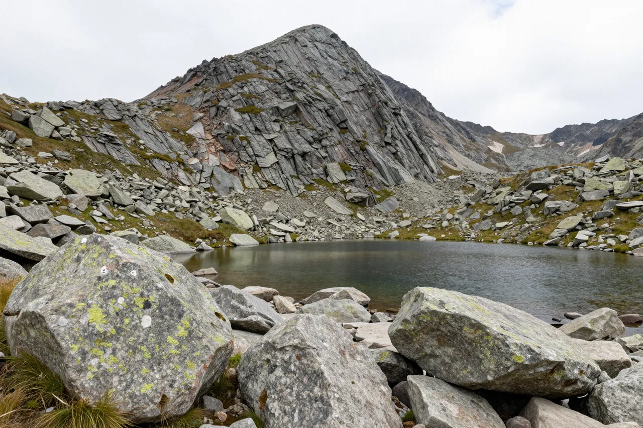 Boulders sit in the foreground of a small mountain tarn under a steep rocky alpine peak