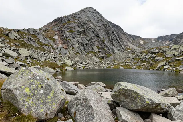 Boulders sit in the foreground of a small mountain tarn under a steep rocky alpine peak