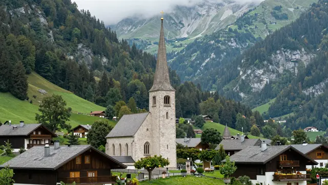 Stone church with a tall steeple sits among wooden chalets in a lush green alpine valley backed by misty mountains