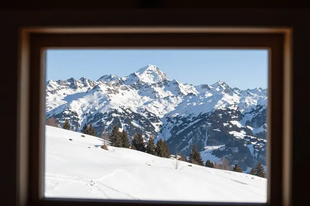 Framed view through a wooden window of sunlit snowy alpine peaks and a pine-dotted snow slope under clear blue sky