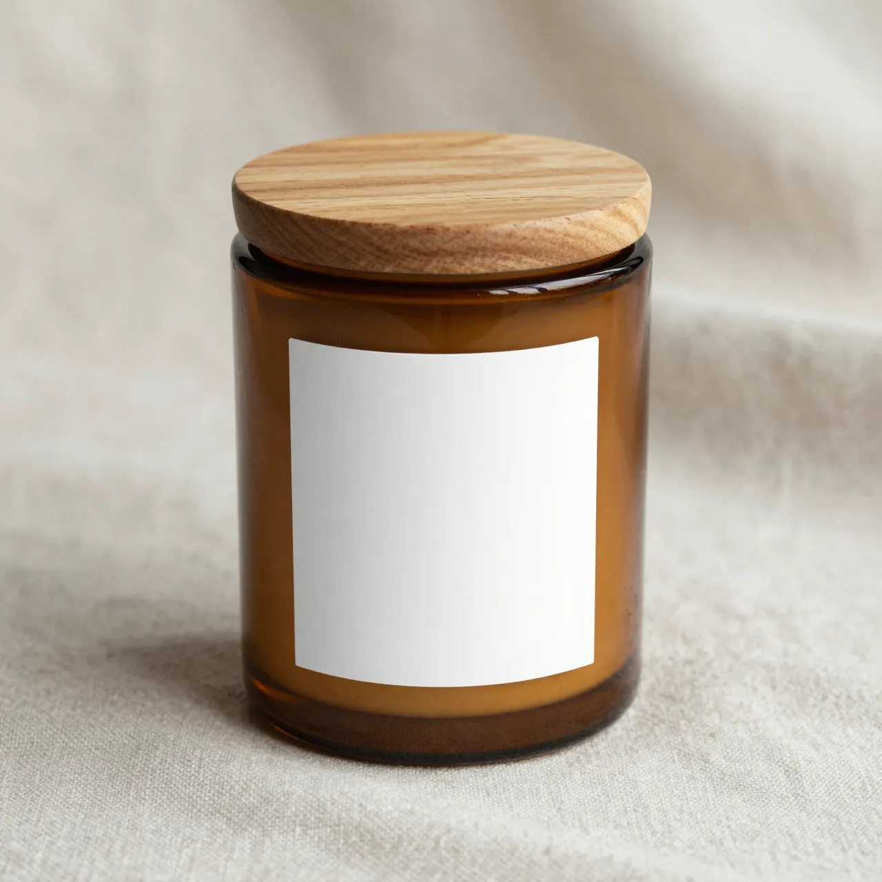 A plain amber glass candle jar with a wooden lid and blank label sits on soft linen cloth