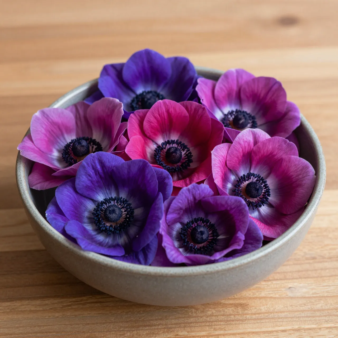 Purple and magenta anemones with dark centers fill a low gray stoneware bowl on a light wooden table
