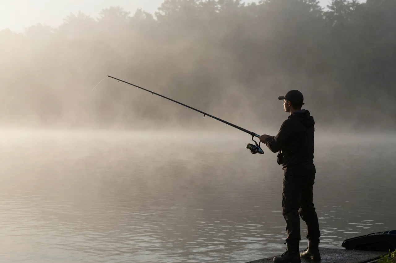 A silhouetted angler stands at the edge of a misty calm lake holding a fishing rod at early dawn.