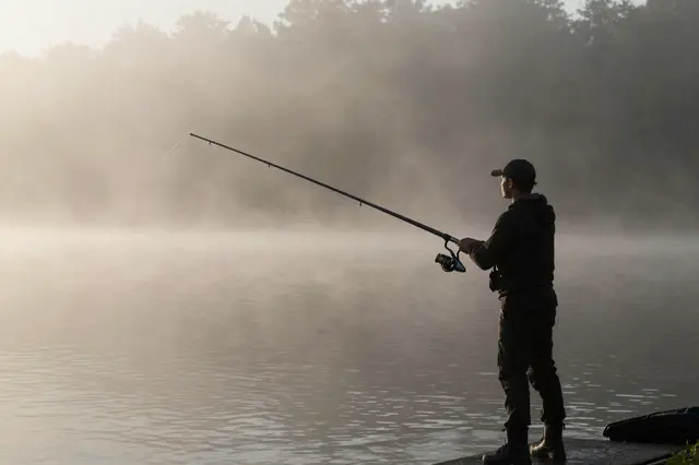 A silhouetted angler stands at the edge of a misty calm lake holding a fishing rod at early dawn.