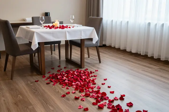Rose petals lead across a wood floor to a candlelit table set for two in a hotel room