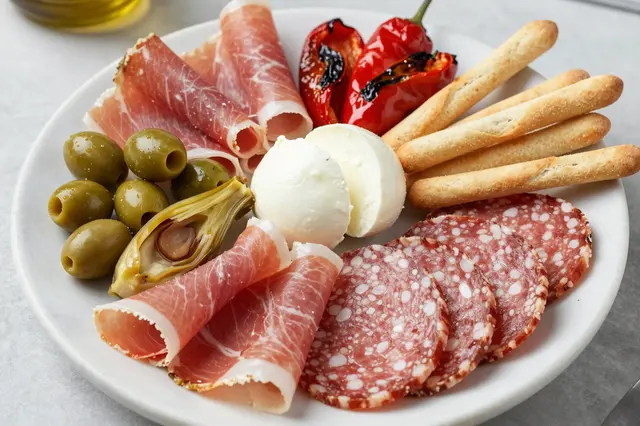 Overhead view of a white plate holding cured meats, mozzarella, olives, roasted peppers, artichoke and breadsticks on a