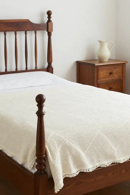 A wooden spindle guest bed with a crocheted coverlet sits next to a wooden nightstand holding a plain ceramic pitcher.