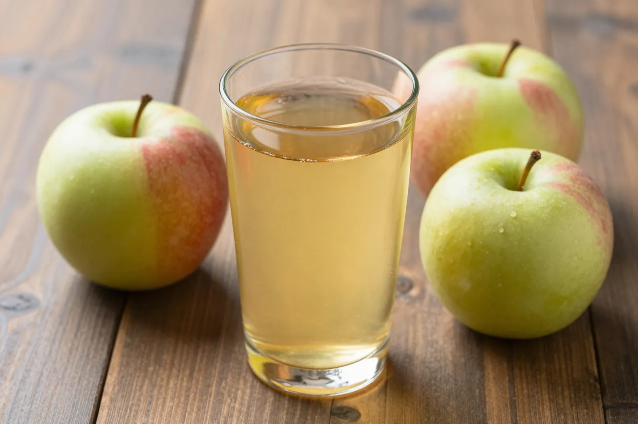 A clear glass of pale apple juice sits surrounded by whole fresh apples on a wooden table