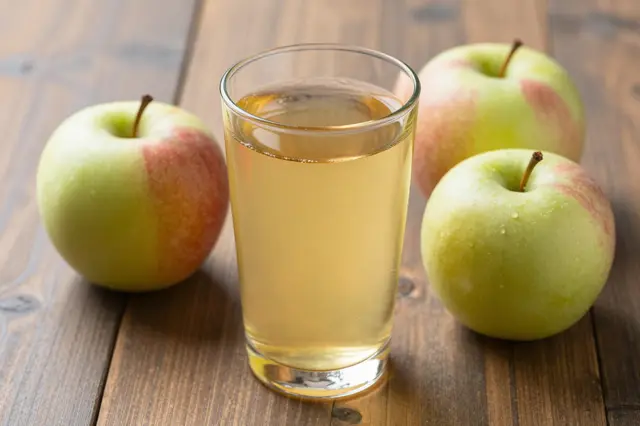 A clear glass of pale apple juice sits surrounded by whole fresh apples on a wooden table