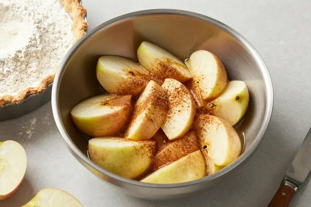 Sliced apples dusted with cinnamon sit in a metal mixing bowl next to an unbaked pie dish on a floured countertop.