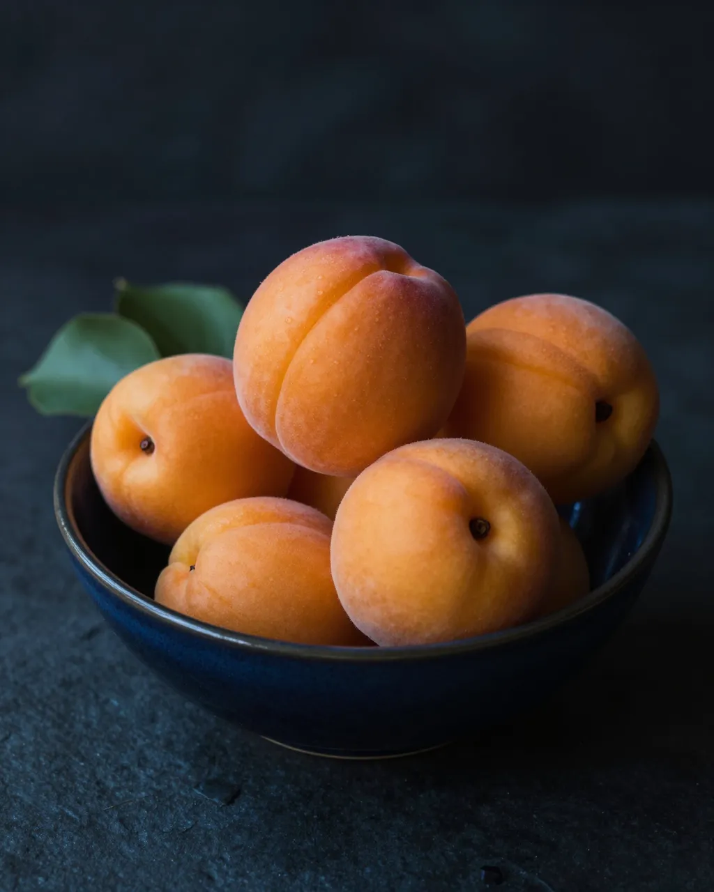 Ripe apricots with green leaves sit in a dark blue bowl against a dark moody background