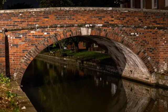 A weathered red brick arched bridge spans a calm still canal after dark